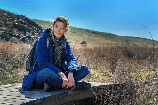 Genç kadın turist oturur ve dağlar ve güneşli gökyüzü arka plan üzerinde eski bir ahşap köprü dayanmaktadır