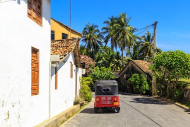 palmiye ağaçları ve bir mavi gökyüzünün ortasında açık güneşli bir günde küçük bir tropikal kasabasında kiremitli çatılı house yakınındaki bir kaldırım taşı kaldırım üzerinde kırmızı tuk-tuk duruyor