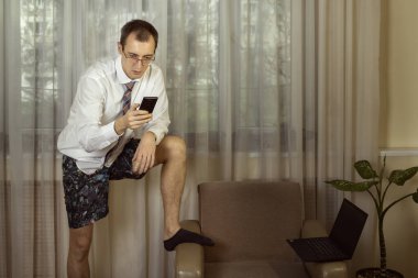 Beyaz gömlekli, kravatlı ve iç çamaşırlı genç adam evde çalışıyor ve akıllı telefondan video konferansı düzenliyor. Renkli. Boşluğu kopyala