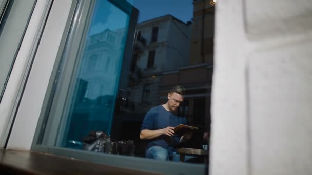 El joven se sienta en la cafetería y trabaja con la tableta — Vídeos de Stock
