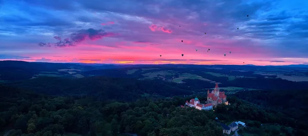 Panorámica, vista aérea del romántico castillo de cuento de hadas Bouzov con globos de aire caliente contra el atardecer rojo — Foto de Stock