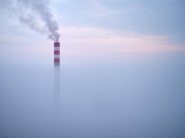 Smoking chimney of a power plant coming out of the inverse fog. The power plant chimney in operation, a red-white striped, rises above the surrounding fog. Energy industry. Heating plant in operation.