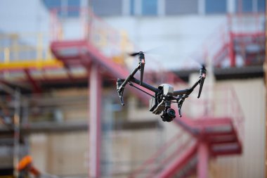 Visual inspection in industry. Controlling of hard to reach objects from the air in the industry. Side view. Hovering  black quadcopter with  powerful camera against blurred industrial construction.
