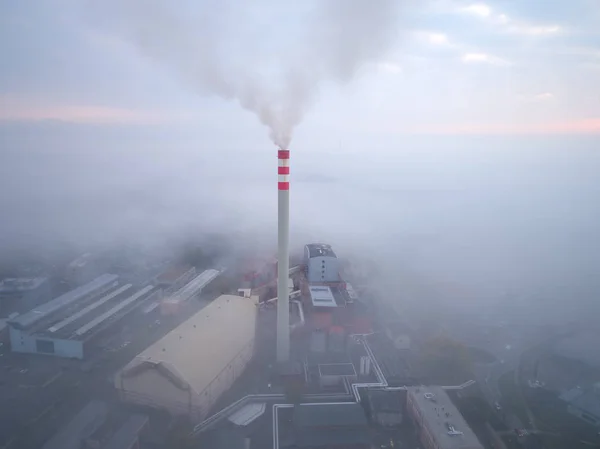 Smoking chimney of a power plant coming out of the inverse fog. The power plant chimney in operation, a red-white striped, rises above the surrounding fog. Energy industry. Heating plant in operation.