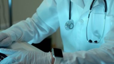 Yaşlı doktor ofisinde dizüstü bilgisayar üzerinde çalışan stetoskop ile karşılaştı. Beyaz önlük, temiz lateks eldiven ve akıllı saat takıyor. Tıp adamı bir reçete yazma veya değiştirme işlemi açıklar