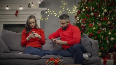 Genç çift kanepede oturan ve akıllı telefon kullanarak arka plan Noel ağacı üzerinde