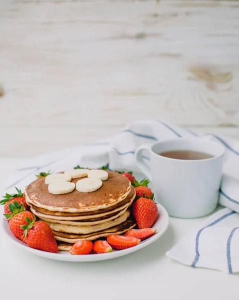 Healthy breakfast. Pancakes with strawberries, bananas, cup of black tea on white background with white towel