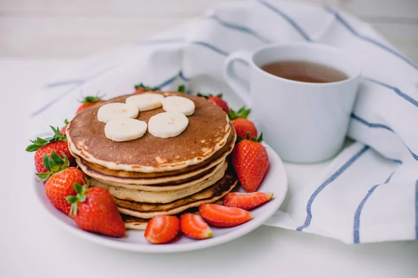 Gesundes Frühstück. Pfannkuchen mit Erdbeeren, Bananen, Tasse schwarzen Tee auf weißem Hintergrund mit weißem Handtuch — Stockfoto