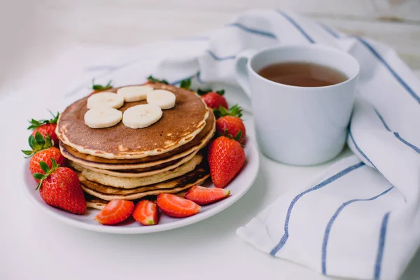 Healthy breakfast. Pancakes with strawberries, bananas, cup of black tea on white background with white towel