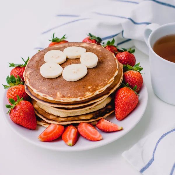 Gezond ontbijt. Pannenkoeken met aardbeien, bananen, kopje zwarte thee op witte achtergrond met witte handdoek — Stockfoto