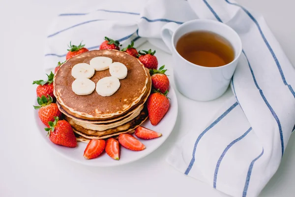 Desayuno saludable. Panqueques con fresas, plátanos, taza de té negro sobre fondo blanco con toalla blanca — Foto de Stock