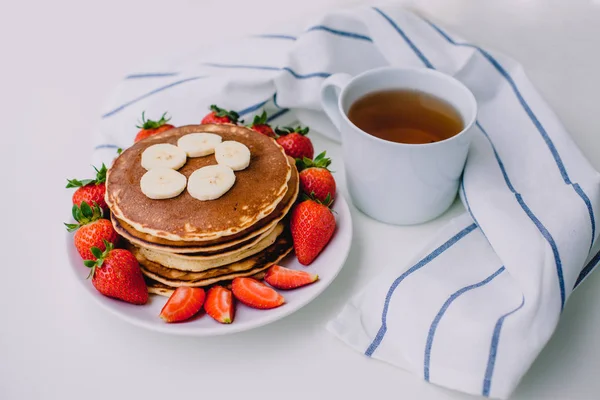 Healthy breakfast. Pancakes with strawberries, bananas, cup of black tea on white background with white towel