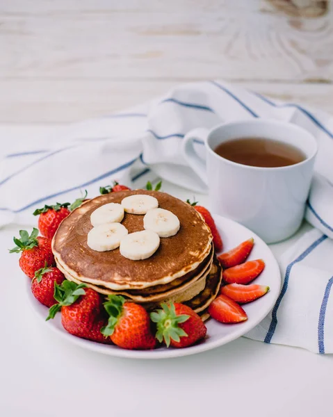 Healthy breakfast. Pancakes with strawberries, bananas, cup of black tea on white background with white towel