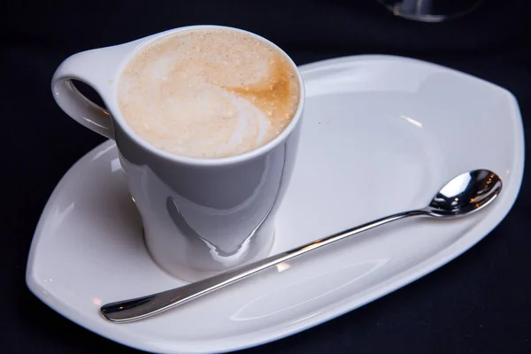 Cup of latte coffee in a retaurant feeding in white cup on white long plate with long latte spoon