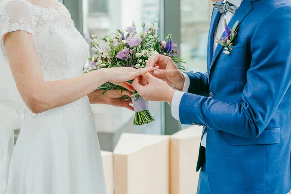 The bride and groom exchange rings. The bride puts the ring on the groom. The groom puts the ring on the bride.