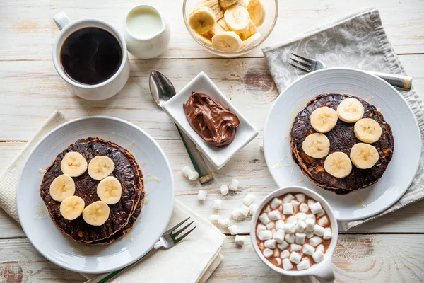 Breakfast for couple included oat pancakes with banana, nutella, honey, with coffe and milk and cacao with marshmallow on white wooden  background