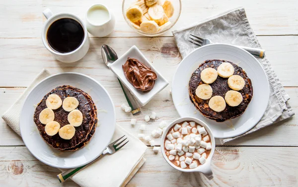 Breakfast for couple included oat pancakes with banana, nutella, honey, with coffe and milk and cacao with marshmallow on white wooden  background