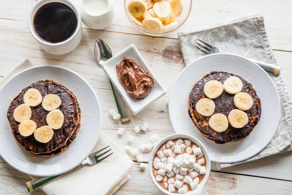 Breakfast for couple included oat pancakes with banana, nutella, honey, with coffe and milk and cacao with marshmallow on white wooden  background