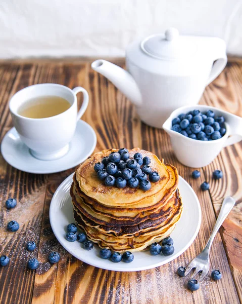 Pancakes healthy breakfast with blueberries, bog whortleberry, cup of green tea, cup of blueberries and teapot on brown wooden background