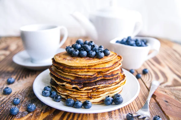 Pancakes healthy breakfast with blueberries, bog whortleberry, cup of green tea, cup of blueberries and teapot on brown wooden background