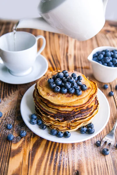 Pancakes healthy breakfast with blueberries, bog whortleberry, cup of green tea, cup of blueberries and teapot on brown wooden background