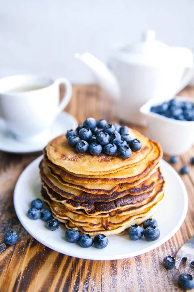 Pancakes healthy breakfast with blueberries, bog whortleberry, cup of green tea, cup of blueberries and teapot on brown wooden background