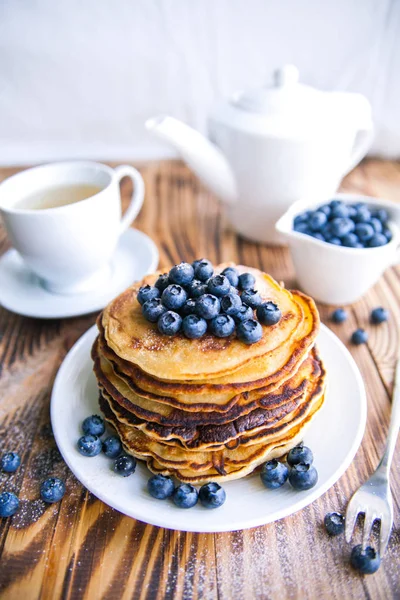 Pancakes healthy breakfast with blueberries, bog whortleberry, cup of green tea, cup of blueberries and teapot on brown wooden background