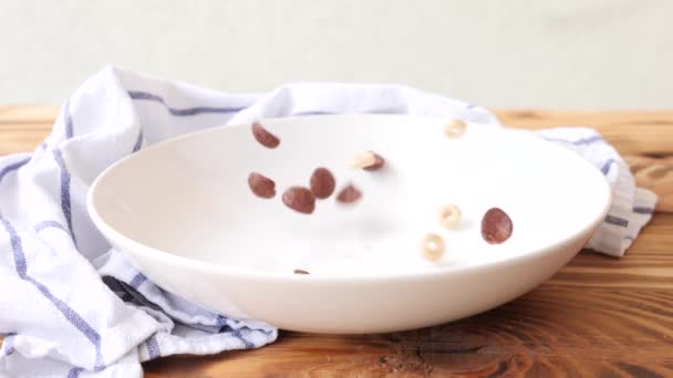 Breakfast included corn flakes, milk and raspberies. Corn flakes are falling on a white plate on wooden background in slow motion — Stock Video