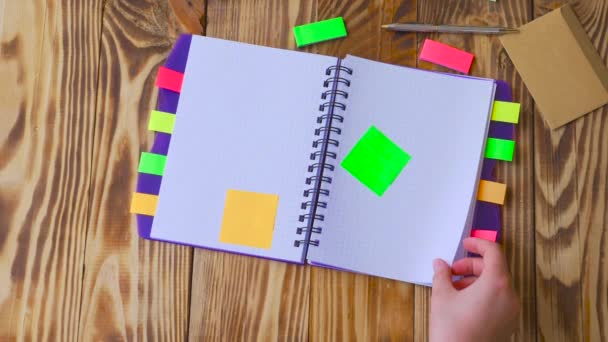 Woman opens notepad and thumbnails pages. Open notepad with office supplies. Open notepad lies on a wooden desktop with marker, pencil, pen and Stickers — Stock Video