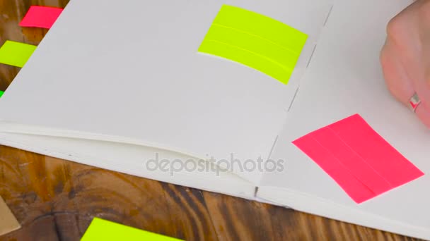 A mulher escreve notas no bloco de notas. Bloco de notas aberto com material de escritório. Bloco de notas aberto está em um desktop de madeira com marcador, lápis, caneta e adesivos — Vídeo de Stock