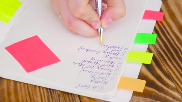Γυναίκα Γράψτε σημειώσεις στο Σημειωματάριο (Notepad). Ανοίξτε το σημειωματάριο με είδη γραφείου. Ανοίξτε το σημειωματάριο βρίσκεται σε μια ξύλινη επιφάνεια εργασίας με μαρκαδόρο, μολύβι, στυλό και αυτοκόλλητα — Αρχείο Βίντεο