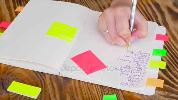 Vrouw notities schrijven in Kladblok. Open Kladblok met kantoorbenodigdheden. Open Kladblok ligt op een houten bureaublad met markeringen, potlood, pen en Stickers — Stockvideo