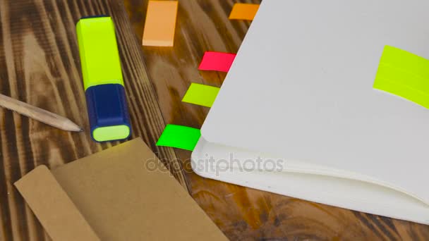 La mujer escribe notas en el bloc de notas. Bloc de notas abierto con material de oficina. Bloc de notas abierto se encuentra en un escritorio de madera con marcador, lápiz, pluma y pegatinas — Vídeos de Stock