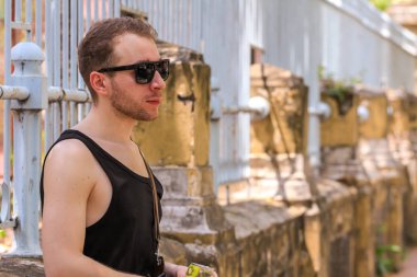 2015.04.26, Bangkok, Tayland. Siyah güneş gözlüğü takan sakallı bir genç çitin kenarında dikiliyor..