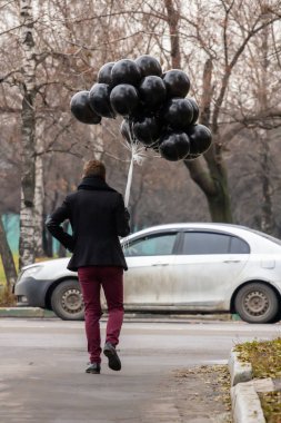 2015.11.22, Moskova, Rusya. Doğum günü hüzünlü bir tatil konseptidir. Siyah eşarp ve ceket giyen genç sarışın bir adam elinde siyah balonlarla caddeden aşağı, arka tarafa doğru gidiyor..