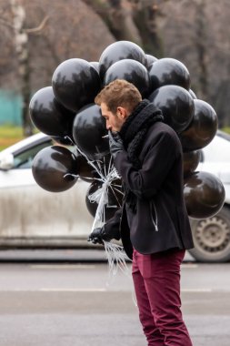 2015.11.22, Moskova, Rusya. Doğum günü hüzünlü bir tatil konseptidir. Siyah atkı ve ceket giyen genç sarışın bir adam elinde siyah balonlar ve sigara içen bir adam..