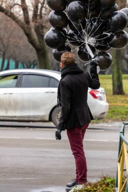 2015.11.22, Moscow, Russia. birthday is a sad holiday concept. A young blonde man wearing black scarf and coat guy holding black balloons and smoking. clipart