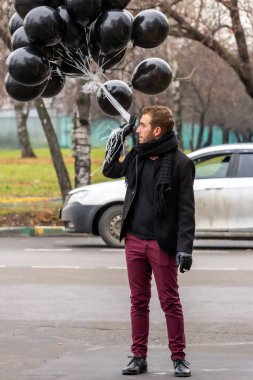 2015.11.22, Moskova, Rusya. Doğum günü hüzünlü bir tatil konseptidir. Siyah atkı ve ceket giyen genç sarışın bir adam elinde siyah balonlar ve sigara içen bir adam..