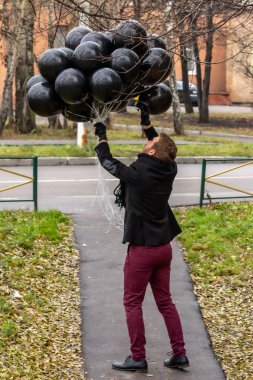 2015.11.22, Moskova, Rusya. Doğum günü hüzünlü bir tatil konseptidir. Siyah atkı ve palto giyen genç sarışın bir adam ağaç dallarına dolanmış düzinelerce balon bırakıyor..