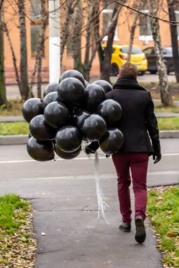 2015.11.22, Moskova, Rusya. Doğum günü hüzünlü bir tatil konseptidir. Siyah eşarp ve ceket giyen genç sarışın bir adam elinde siyah balonlarla caddeden aşağı, arka tarafa doğru gidiyor..