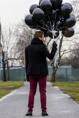 2015.11.22, Moskova, Rusya. Doğum günü hüzünlü bir tatil konseptidir. Siyah eşarp ve ceket giyen genç sarışın bir adam elinde siyah balonlarla caddeden aşağı, arka tarafa doğru gidiyor..