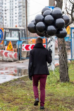 2015.11.22, Moskova, Rusya. Doğum günü hüzünlü bir tatil konseptidir. Siyah eşarp ve ceket giyen genç bir adam şehir manzarası, bulanıklık ve tahıl efekti arka planında siyah balonlar tutuyor..