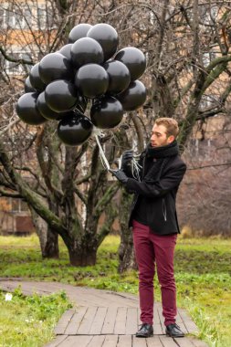 2015.11.22, Moskova, Rusya. Doğum günü hüzünlü bir tatil konseptidir. Siyah eşarp ve palto giyen genç sarışın bir adam elma ağaçlarının arka planında duran siyah balonlar tutuyor..
