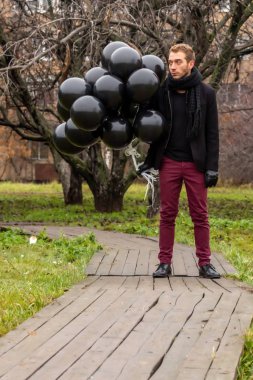2015.11.22, Moskova, Rusya. Doğum günü hüzünlü bir tatil konseptidir. Siyah eşarp ve palto giyen genç sarışın bir adam elma ağaçlarının arka planında duran siyah balonlar tutuyor..
