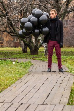 2015.11.22, Moskova, Rusya. Doğum günü hüzünlü bir tatil konseptidir. Siyah eşarp ve palto giyen genç sarışın bir adam elma ağaçlarının arka planında duran siyah balonlar tutuyor..