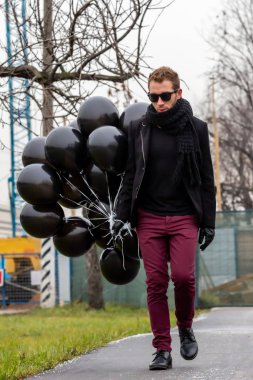 2015.11.22, Moskova, Rusya. Doğum günü hüzünlü bir tatil konseptidir. Siyah fularlı ve ceketli genç sarışın bir adam elinde siyah balonlarla caddeden aşağı doğru gidiyor..