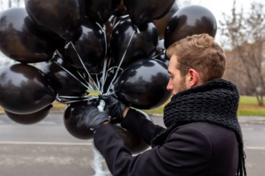 2015.11.22, Moskova, Rusya. Doğum günü hüzünlü bir tatil konseptidir. Siyah atkı ve ceket giyen genç sarışın bir adam elinde siyah balonlar ve başka yöne bakıyor..