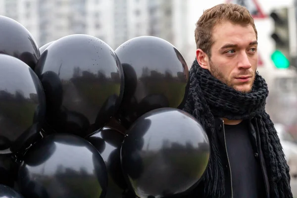 2015.11.22, Moscow, Russia. birthday is a sad holiday concept. A young blonde man wearing black scarf and coat holding black balloons and looking away.