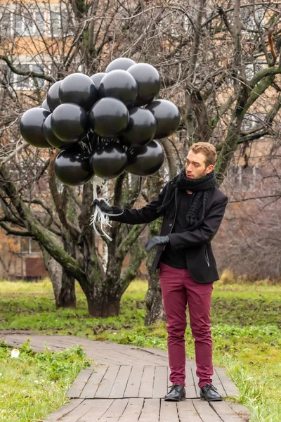 2015.11.22, Moscow, Russia. birthday is a sad holiday concept. A young blonde man wearing black scarf and coat holding black balloons standing on background of apple trees garden.