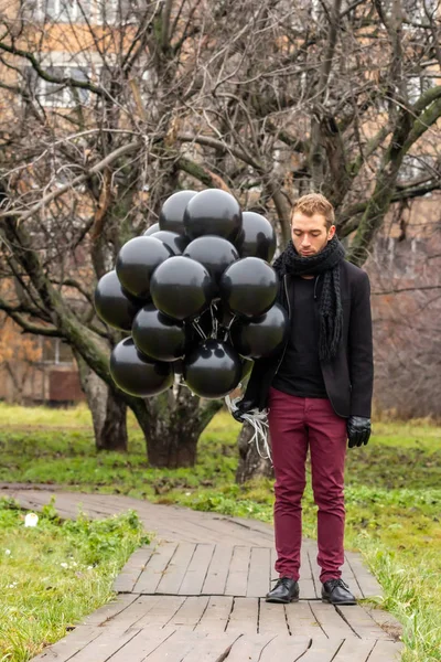 2015.11.22, Moscow, Russia. birthday is a sad holiday concept. A young blonde man wearing black scarf and coat holding black balloons standing on background of apple trees garden.